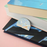 Semi-circular, lilac pin with the words Books Are Magic in capital letters. There is an open book at the bottom and many magical items are bursting forth from the pages, including a wand, raven skull, quill, stars and bubbles. The pin is leaning against a stack of books.