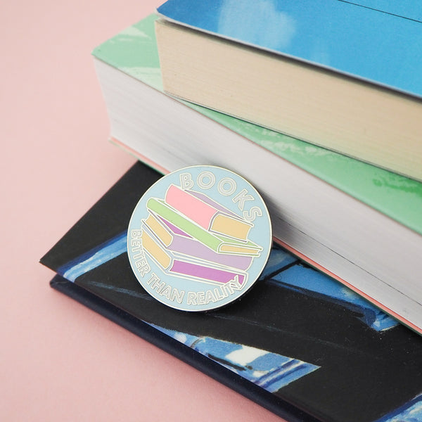 Light blue, circular enamel pin showing a stack of pastel-coloured books in the centre. Around the edge are the words Books Better Than Reality in white capital letters. The pin is leaning against a stack of books.