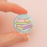 Light blue, circular enamel pin showing a stack of pastel-coloured books in the centre. Around the edge are the words Books Better Than Reality in white capital letters. The pin is being held between a forefinger and thumb.