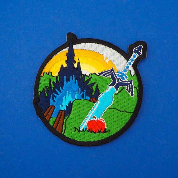 Embroidered patch featuring a sword and apple in the foreground with a castle and sunrise in the background. Patch has a black merrowed edge.