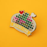 Enamel pin depicting a pixelated image on a plate with a red heart and bone protruding. A banner on the bottom reads Dubious Cook. Pin is on a yellow background.