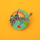 Enamel pin featuring a sword and apple in the foreground with a castle and sunrise in the background. The pin is on a yellow background.