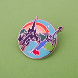 Enamel pin featuring a sword and apple in the foreground with a castle and sunrise in the background. The pin is on a green background.