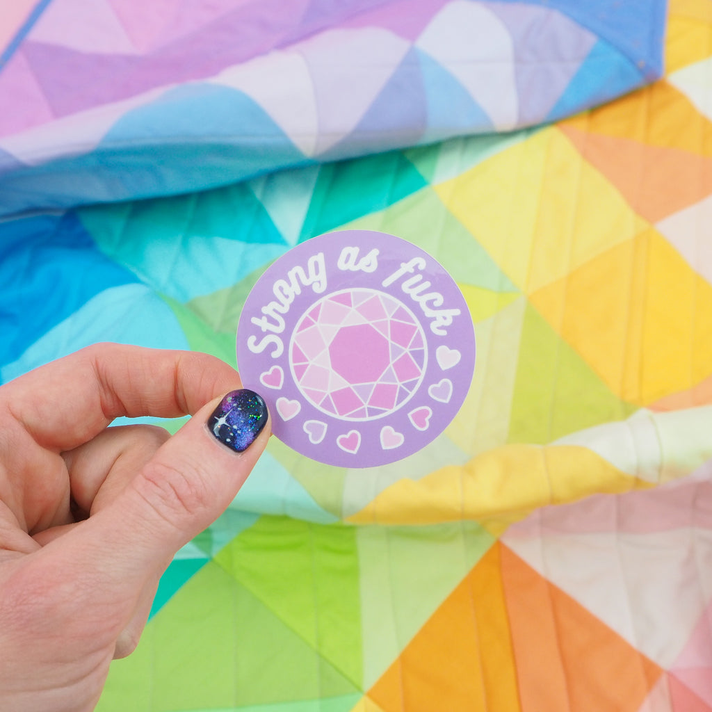Round, purple vinyl sticker. It has a multi-faceted pink gem in the centre. At the top are the words Strong as fuck and below the gem are small hearts in shades of pink. It is held between a finger and thumb.