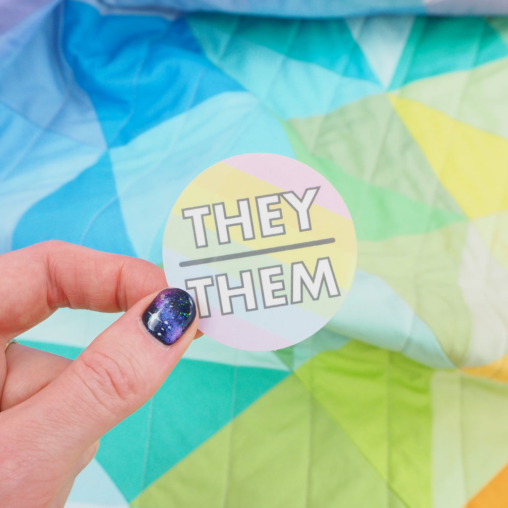 Round vinyl sticker with pastel rainbow diagonal stripes. There is a short horizontal line in the centre; above it is the word They and below it, Them. The lettering is white block capitals. It is held between a finger and thumb.