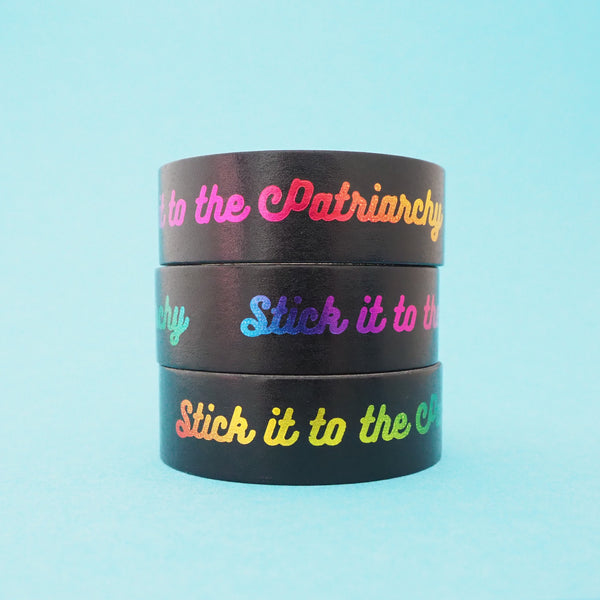 A stack of black washi tape that reads Stick it to the Patriarchy in a gradient rainbow foil.