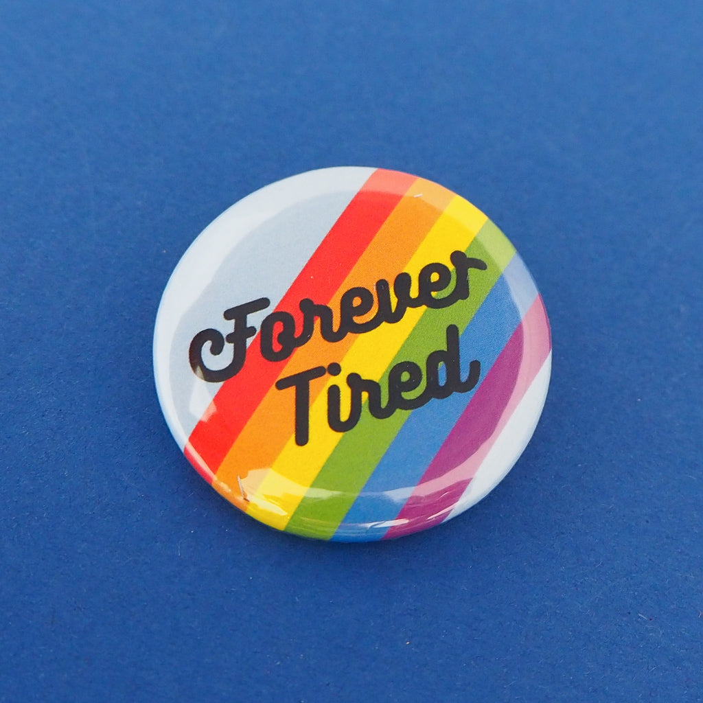 Round button badge. It reads Forever Tired in black script on a pale blue background with diagonal rainbow stripes.