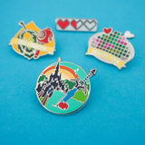 Four enamel pins. The pin in the foreground features a sword and apple in the foreground with a castle and sunrise in the background. The three pins in the background are out of focus.