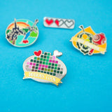 Four Enamel pins. The pin in the foreground depicts a pixelated image on a plate with a red heart and bone protruding. A banner on the bottom reads Dubious Cook. The three pins in the background are out of focus.