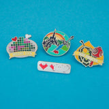Four enamel pins. The first pin is showing three pixelated hearts. The second pin features a sword and apple in the foreground with a castle and sunrise in the background. The third pin shows a pixelated image on a plate with a heart and bone showing. A banner on the bottom reads Dubious Cook. The fourth pin features a leaf and red maraca on a korok seed background, a banner at the bottom reads Seed Collector.