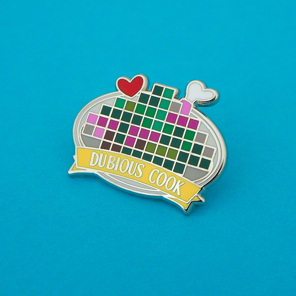 Enamel pin depicting a pixelated image on a plate with a red heart and bone protruding. A banner on the bottom reads Dubious Cook. The pin is on a blue background.