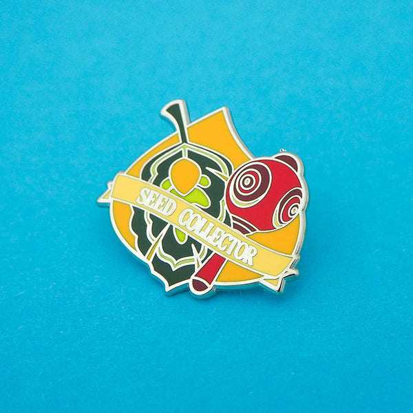 Enamel pin featuring a green leaf and red maraca on a korok seed background, a banner at the bottom reads Seed Collector. The pin is on a blue background.