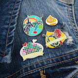 Four pins on a denim jacket. The first enamel pin features a sword and apple in the foreground with a castle and sunrise in the background. The second is a small, glittery korok seed. The third shows a pixelated image on a plate with a heart and bone showing. A banner on the bottom reads Dubious Cook. The fourth pin features a leaf and red maraca on a korok seed background, a banner at the bottom reads Seed Collector.