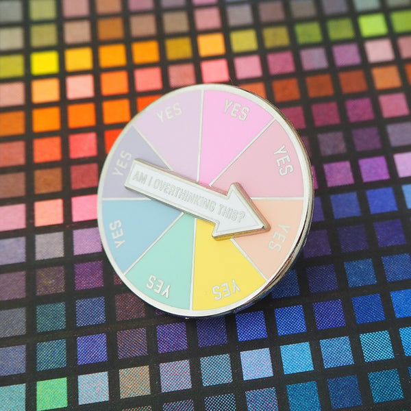 Circular enamel pin divided into eight pastel coloured sections. Each section has the word Yes in capital letters. There is a white arrow that can spin to point to any section with the words Am I Overthinking This? Pin is on a multicoloured grid background.