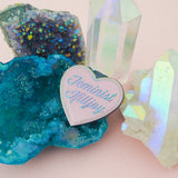 Pastel pink enamel pin in the shape of a heart. The words Feminist Killjoy are written in a flowing script and are rainbow-plated. Rainbow-plated dots run along the inside border of the heart. An epoxy resin coating gives the pin a shiny finish. The pin is resting on a cluster of coloured crystals on a pale pink background.