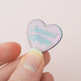 Pastel pink enamel pin in the shape of a heart. The words Feminist Killjoy are written in a flowing script and are rainbow-plated. Rainbow-plated dots run along the inside border of the heart. An epoxy resin coating gives the pin a shiny finish. The pin is being held between a finger and thumb in front of a pale pink backdrop.