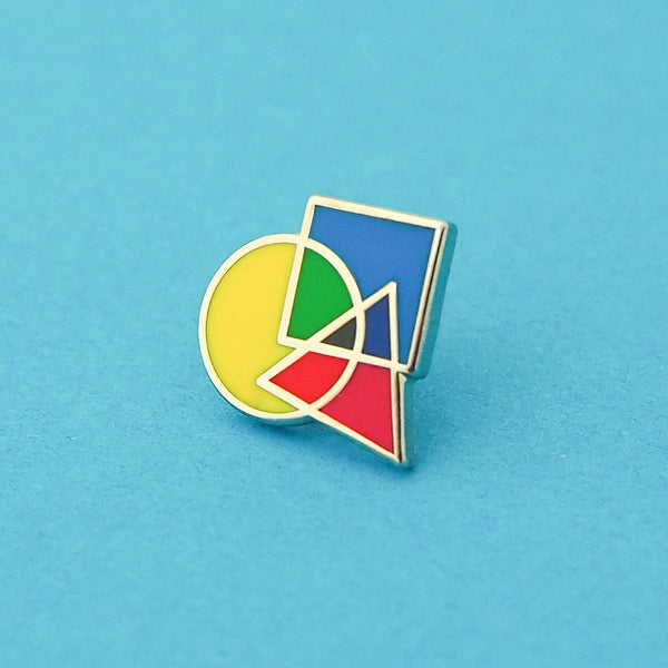 Enamel pin featuring an overlapping circle, square, and triangle. The shapes are brightly coloured, with the colours mixing where they overlap. Pin is shown on a light blue background.