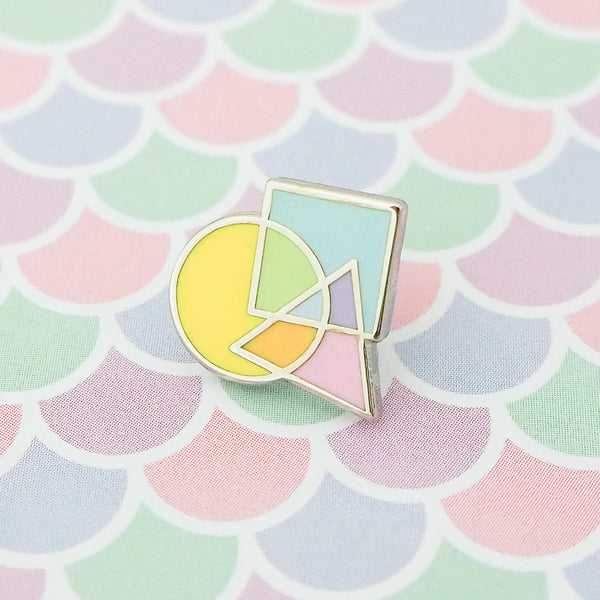 Enamel pin featuring an overlapping circle, square, and triangle. The shapes are pastel coloured, with the colours mixing where they overlap. The pin is shown on a pastel, fish-scale patterned background.