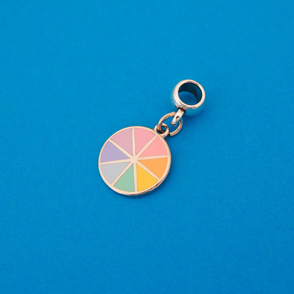 Enamel charm on a smooth cup bail bead. Charm is circular and split into eight sections. Each section is filled with a pastel rainbow colour, replicating a colour wheel.
