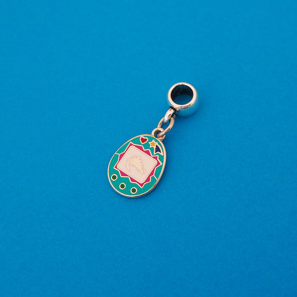 Enamel charm on a smooth cup bail bead. Charm is a teal, egg-shaped virtual pet toy. On the white screen is a silver stegosaur and game icons. Beneath the screen are 3 green buttons and above are a heart and shooting star in bright colours.