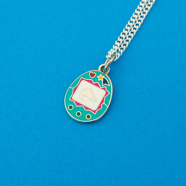 Silver-plated curb chain necklace with an enamel charm pendant. Charm is a teal, egg-shaped virtual pet toy. On the white screen is a silver stegosaur and game icons. Beneath the screen are 3 green buttons and above are a heart and shooting star in bright colours