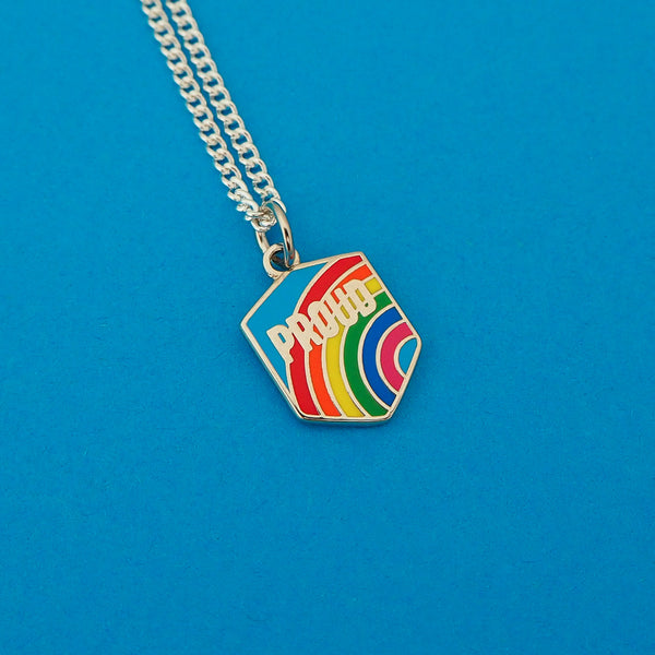 Silver-plated curb chain necklace with an enamel charm pendant. Charm is in a shield shape and reads Proud in silver capital letters. The background is a bright rainbow on a blue sky.