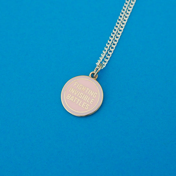Silver-plated curb chain necklace with an enamel charm pendant. Charm is round and pastel pink. It bears the words Fighting Invisible Battles in silver capitals.