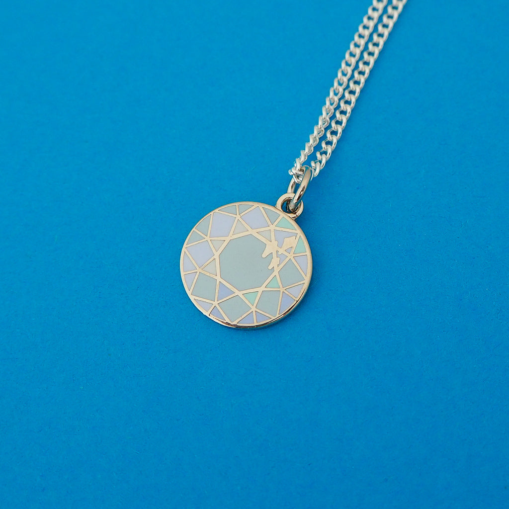 Silver curb-chain necklace with an enamel charm pendant. Charm is circular and depicts a blue, multi-faceted gem.