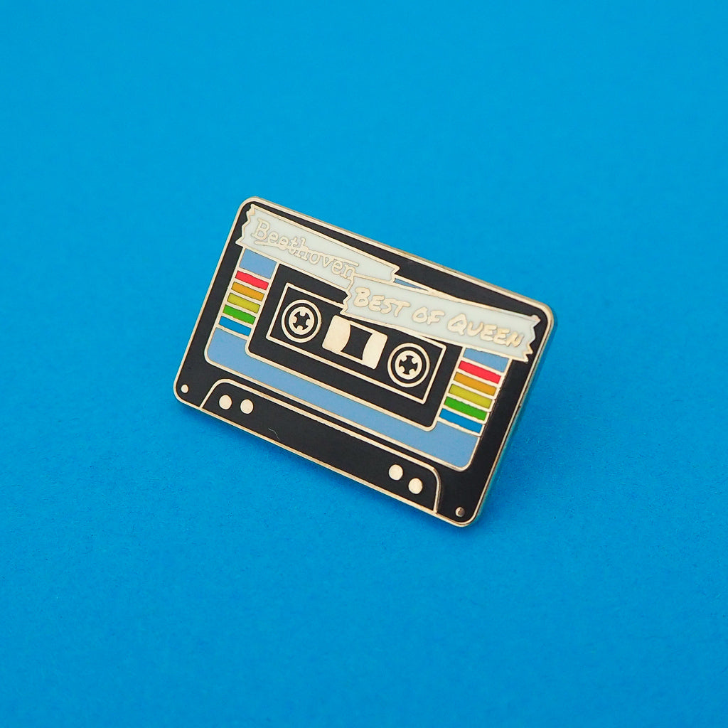 Enamel pin depicting a black cassette tape. The centre of the tape is blue with a rainbow stripe. At the top the word Beethoven has been scored out and the words Best of Queen are written in capital letters. Pin is shown on a blue background.