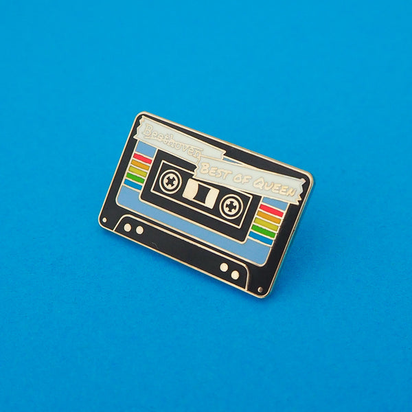 Enamel pin depicting a black cassette tape. The centre of the tape is blue with a rainbow stripe. At the top the word Beethoven has been scored out and the words Best of Queen are written in capital letters. Pin is shown on a blue background.