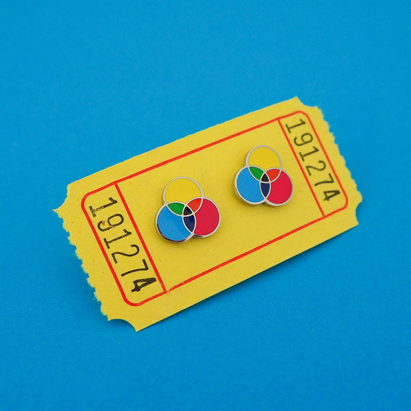 Metal stud earrings with three overlapping circles. The circles are filled cyan, magenta, and yellow enamel, with the colours mixing in the areas where they overlap. The earrings are on a yellow carnival ticket.