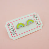 A pair of enamel stud earrings depicting pastel coloured rainbows. The earrings are on a white carnival ticket.
