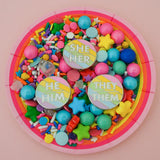 Three round enamel pins sitting on a plate of candy. Each pin has pastel rainbow diagonal stripes. There is a short horizontal line in the centre with pronouns above and below. They read individually in white capitals; She/Her, He/Him, or They/Them.