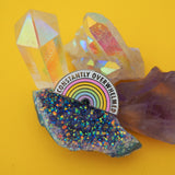 Shaped enamel pin depicting a pastel rainbow. Above the rainbow is a band of white where the words Constantly Overwhelmed are written in capital letters.  The pin is nestled amongst a cluster of colourful crystals.