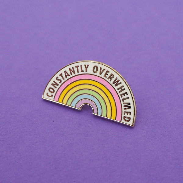 Shaped enamel pin depicting a pastel rainbow. Above the rainbow is a band of white where the words Constantly Overwhelmed are written in capital letters. The pin is on a purple background.