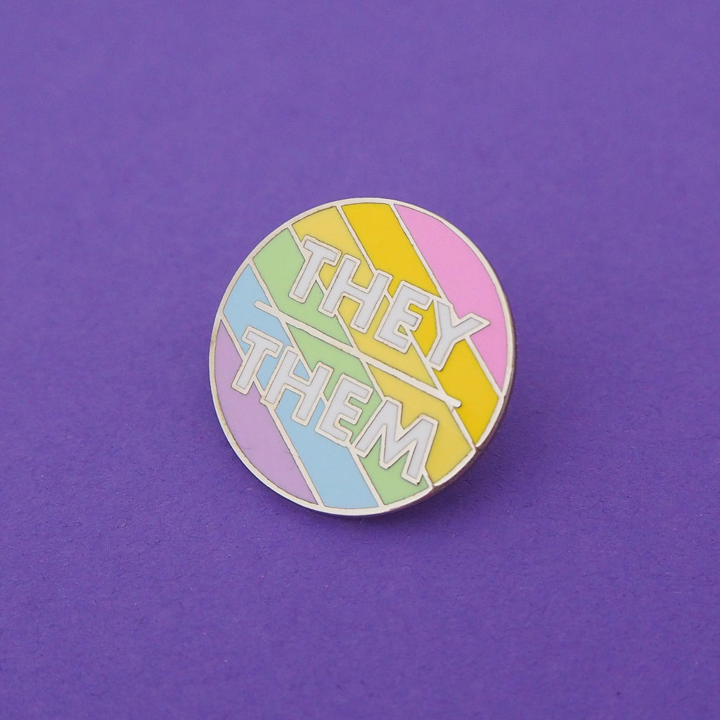 Round enamel pin with pastel rainbow diagonal stripes. There is a short horizontal line in the centre; above it is the word They and below it, Them. The lettering is white block capitals. The pin is on a deep purple background.