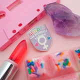 Pink enamel pin in a shield shape. At the top it reads Behold My Feminist Agenda. The word Behold is filled in rainbow colours. At the bottom there is a feminine symbol with a pink heart to the left and a blue heart to the right. The pin is on a pink background surrounded by a lipstick, cassette, chocolate, and a crystal.