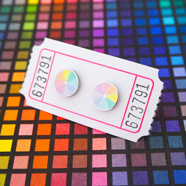 A pair of enamel stud earrings. The earrings are circular and split into eight sections. Each section is filled with a pastel rainbow colour, replicating a colour wheel. The earrings are on a white carnival ticket.