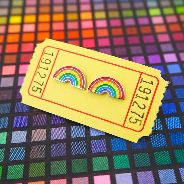 A pair of enamel stud earrings depicting brightly coloured rainbows. The earrings are on a yellow carnival ticket.