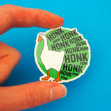 Green, circular vinyl sticker. On the left is a goose and the rest of the sticker is filled with the word Honk repeated in different sizes in black capitals. The goose and letters protrude over the edge of the circle. The sticker has a white border and is held between a finger and thumb.