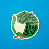 Green, circular vinyl sticker. On the left is a goose and the rest of the sticker is filled with the word Honk repeated in different sizes in black capitals. The goose and letters protrude over the edge of the circle. The sticker has a white border.