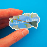 Vinyl sticker showing an oval lake with stones and weeds around its edge. Across the lake is a large green rake and the words Rake in the Lake in white script. The sticker has a white border and is held between a finger and thumb.