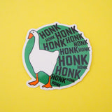 Green, circular vinyl sticker. On the left is a goose and the rest of the sticker is filled with the word Honk repeated in different sizes in black capitals. The goose and letters protrude over the edge of the circle. The sticker has a white border.