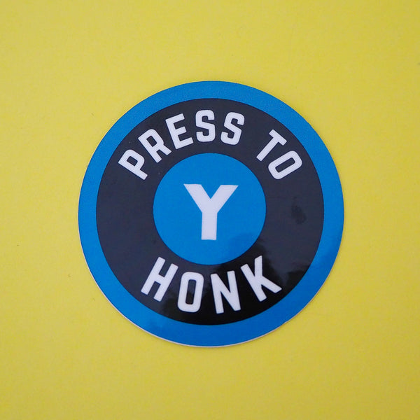 Round vinyl sticker that reads Press To Honk in white capitals within a black ring. A blue dot is in the middle of the black ring and has a white capital Y in the centre, mimicking a gaming controller button. The sticker has a blue border.
