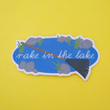 Vinyl sticker showing an oval lake with stones and weeds around its edge. Across the lake is a large green rake and the words Rake in the Lake in white script. The sticker has a white border.