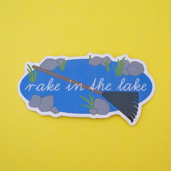 Vinyl sticker showing an oval lake with stones and weeds around its edge. Across the lake is a large green rake and the words Rake in the Lake in white script. The sticker has a white border.