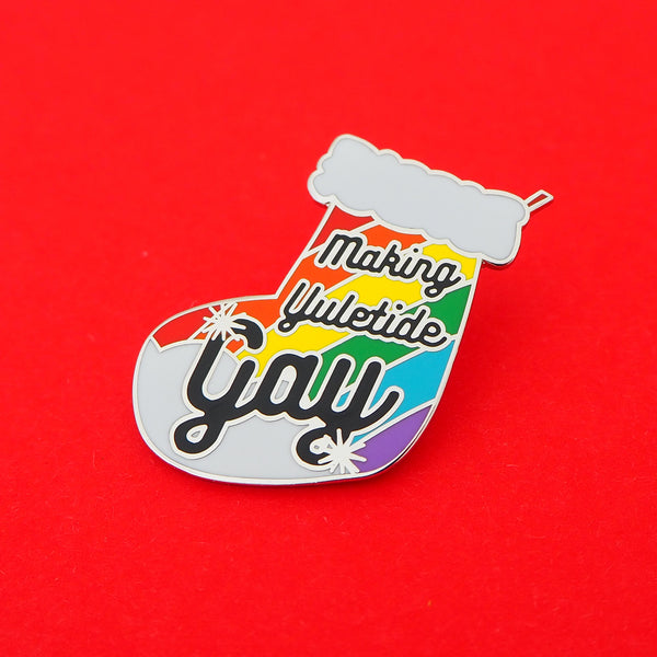 Enamel pin in the shape of a Christmas stocking with rainbow stripes, it reads Making Yuletide Gay in black script. The word Gay is larger and has star sparkles. The pin is on a bright red background.