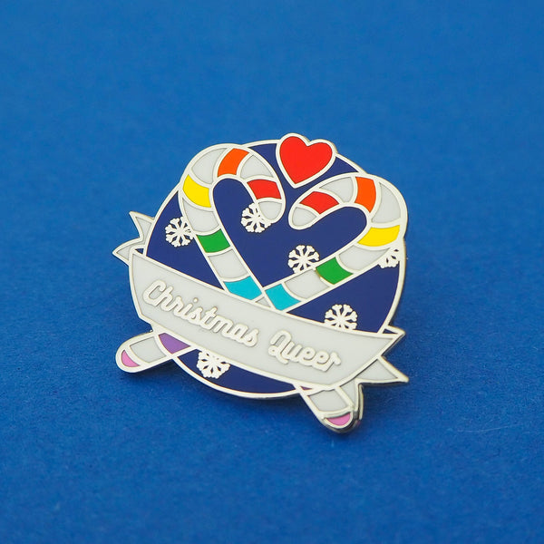 Round enamel pin showing two rainbow striped candy canes crossed at an angle which makes their hooks form a heart shape. There is a small red heart above them and the background is snowflakes on dark blue. There is a ribbon banner at the bottom which reads Christmas Queer. The pin is shown on a blue background.