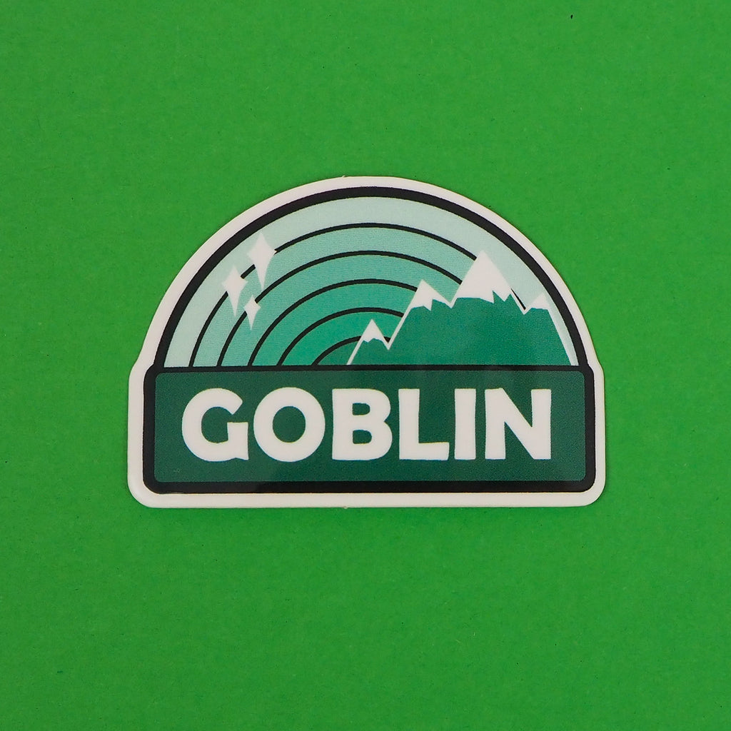 Vinyl sticker with a green ombre rainbow above a dark green banner which reads Goblin in white capital letters. On the right of the rainbow are green mountains with silver caps, and the left has white sparkles. The sticker has a white border.