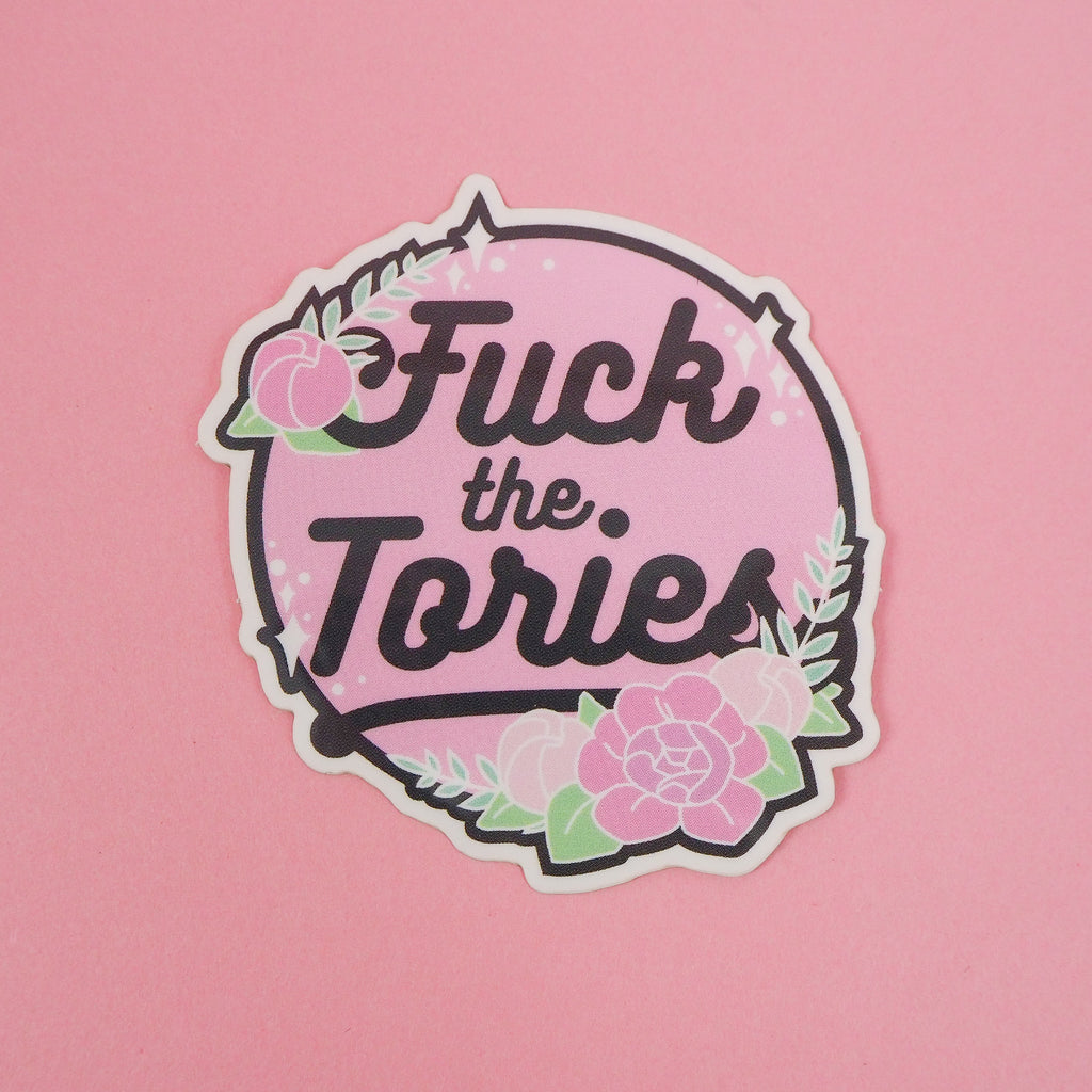 Circular, pastel pink vinyl sticker with the words Fuck the Tories in a black script. The sticker is decorated with pink roses, green foliage and white sparkles, all bursting over the edge of the circle. The sticker has a white edge.